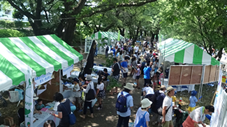 台田運動公園内の陸の会場。桜並木の公園で、木陰の中に展示ブースが設置され、緑陰が涼しい。