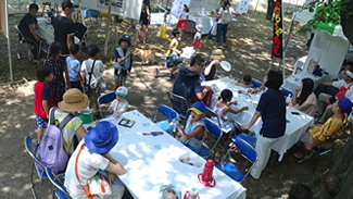 台田運動公園内の陸の会場。桜並木の公園で、木陰の中に展示ブースが設置され、緑陰が涼しい。