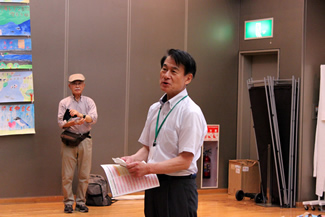イベントが終了し、来場者が帰っていったあと、出展者を含む関係者全員を前に、簡単な総括をする環境フェスティバル実行委員長の柘植正憲さん。手作り体験広場の机などを撤収して組み上げたホール内の雛壇に参加者が並んで記念撮影を撮るのが恒例になっている。