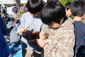 校庭に出てまずしたのは、自分たちが育てたお米の籾摺り。子どもたちは、それぞれ自宅からすり鉢や網かごなどを持ってきて、擂り粉木（すりこぎ）で擦りながら籾を外す。ある程度外れてきたら、息を吹きかけると軽い籾殻が飛んでいって、手元に玄米だけが残る。