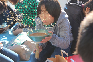 校庭に出てまずしたのは、自分たちが育てたお米の籾摺り。子どもたちは、それぞれ自宅からすり鉢や網かごなどを持ってきて、擂り粉木（すりこぎ）で擦りながら籾を外す。ある程度外れてきたら、息を吹きかけると軽い籾殻が飛んでいって、手元に玄米だけが残る。