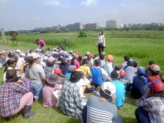 多摩川と大栗川の合流地点の土手の上にて、講師の羽澄ゆり子さんに挨拶をする。この後、土手下の河原から棚橋校長も登場する。