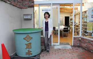 雨水市民の会の副理事長・高橋朝子さん。鳩の街商店街にある雨水市民の会事務所の前にて。玄関前に置かれた雨水タンク（天水桶）は、嘉永六年鋳造、ちょうど黒船来航に沸いた年だ。先祖代々受け継がれてきた天水桶が、関東大震災や東京大空襲などにより、引っ越し先に移設されていたものを、誕生地である墨田に里帰りさせたいと相談があって寄贈された。2015年5月に天水桶里帰りプロジェクトと銘打って移設作業を行い、錆落としと再塗装を行った。現在は、雨水市民の会事務所前に鎮座している。