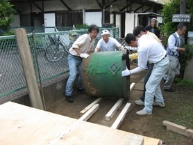 雨水市民の会の副理事長・高橋朝子さん。鳩の街商店街にある雨水市民の会事務所の前にて。玄関前に置かれた雨水タンク（天水桶）は、嘉永六年鋳造、ちょうど黒船来航に沸いた年だ。先祖代々受け継がれてきた天水桶が、関東大震災や東京大空襲などにより、引っ越し先に移設されていたものを、誕生地である墨田に里帰りさせたいと相談があって寄贈された。2015年5月に天水桶里帰りプロジェクトと銘打って移設作業を行い、錆落としと再塗装を行った。現在は、雨水市民の会事務所前に鎮座している。