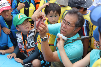 子どもたちに囲まれながら、魚の捌き方から串刺しの仕方まで実演して説明をする、講師の山ちゃん。見つめる子どもたちは真剣な眼差しだ。